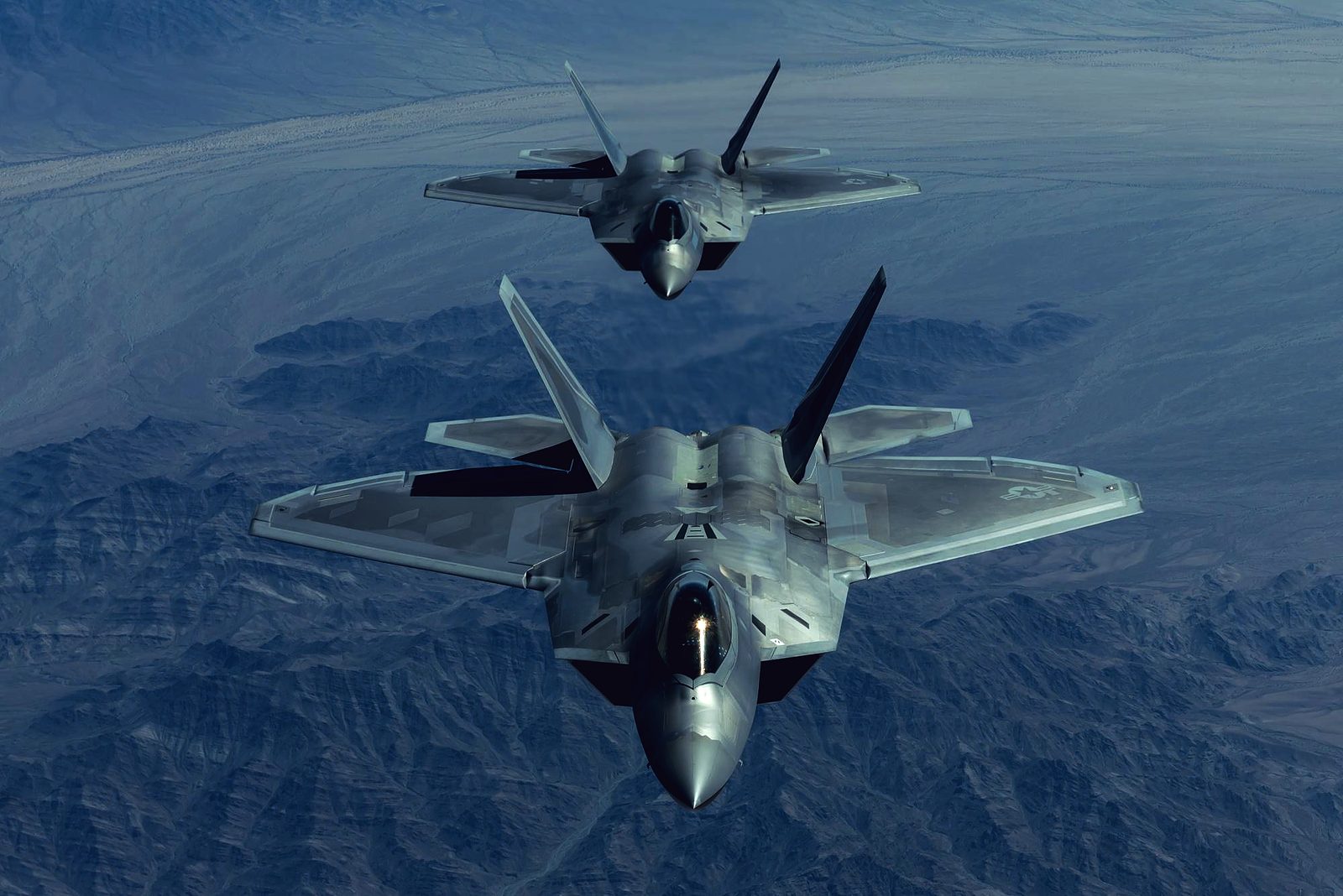 Two U.S. Air Force F-22 Raptors fly in formation toward the camera, high above desert mountains.